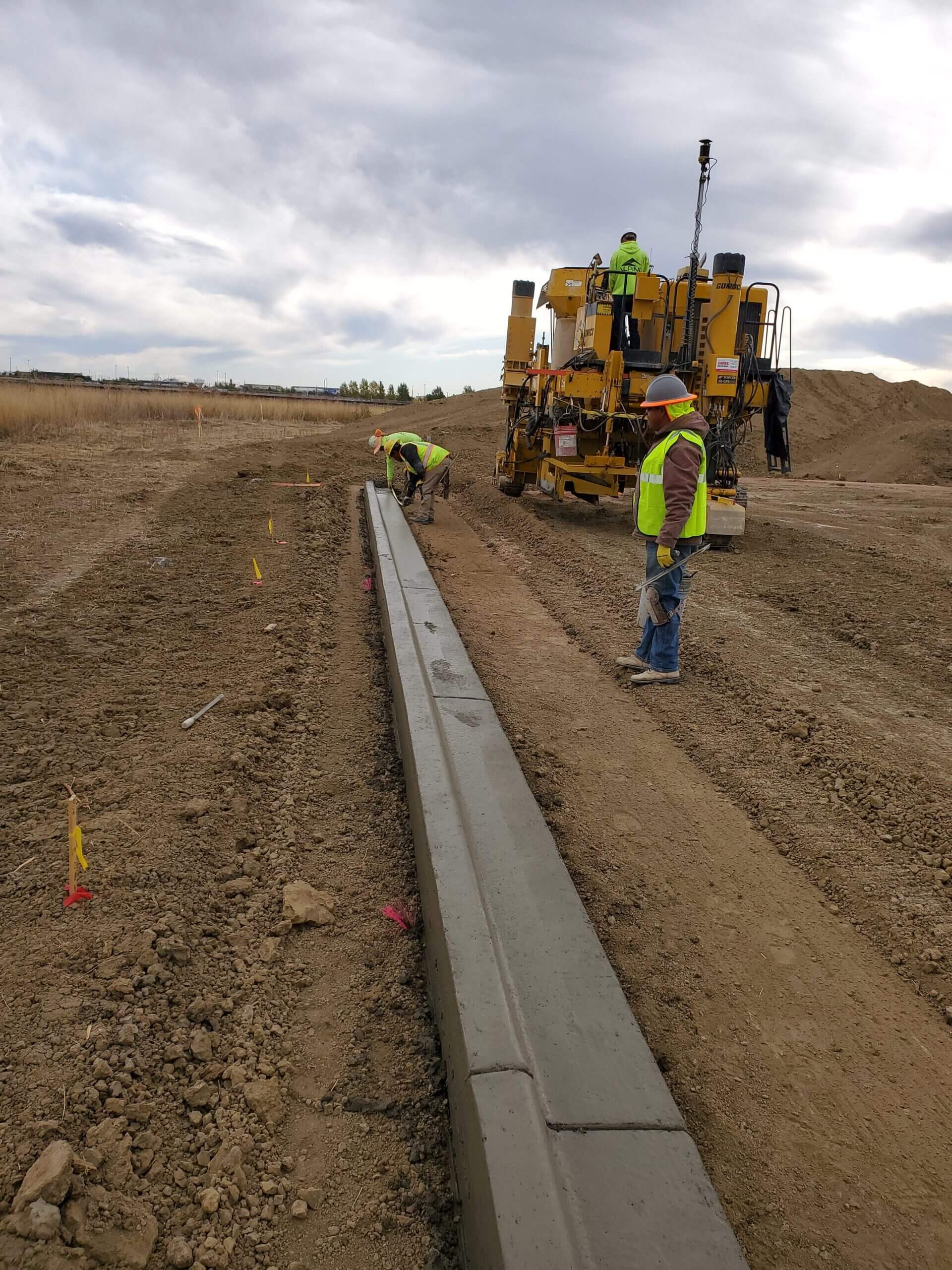 Construction workers installing concrete curb and gutter using a curb machine at an active roadway development project.