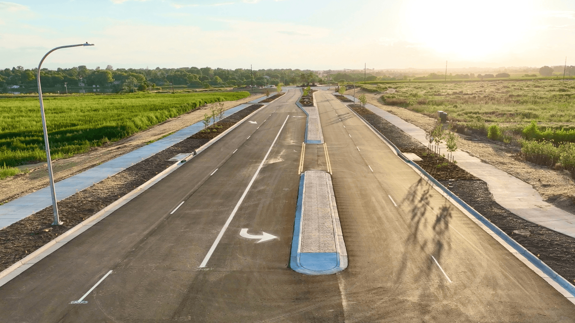 Wide view of a newly constructed divided roadway with landscaped median islands, sidewalks, and streetlights, running through open green fields during sunset.