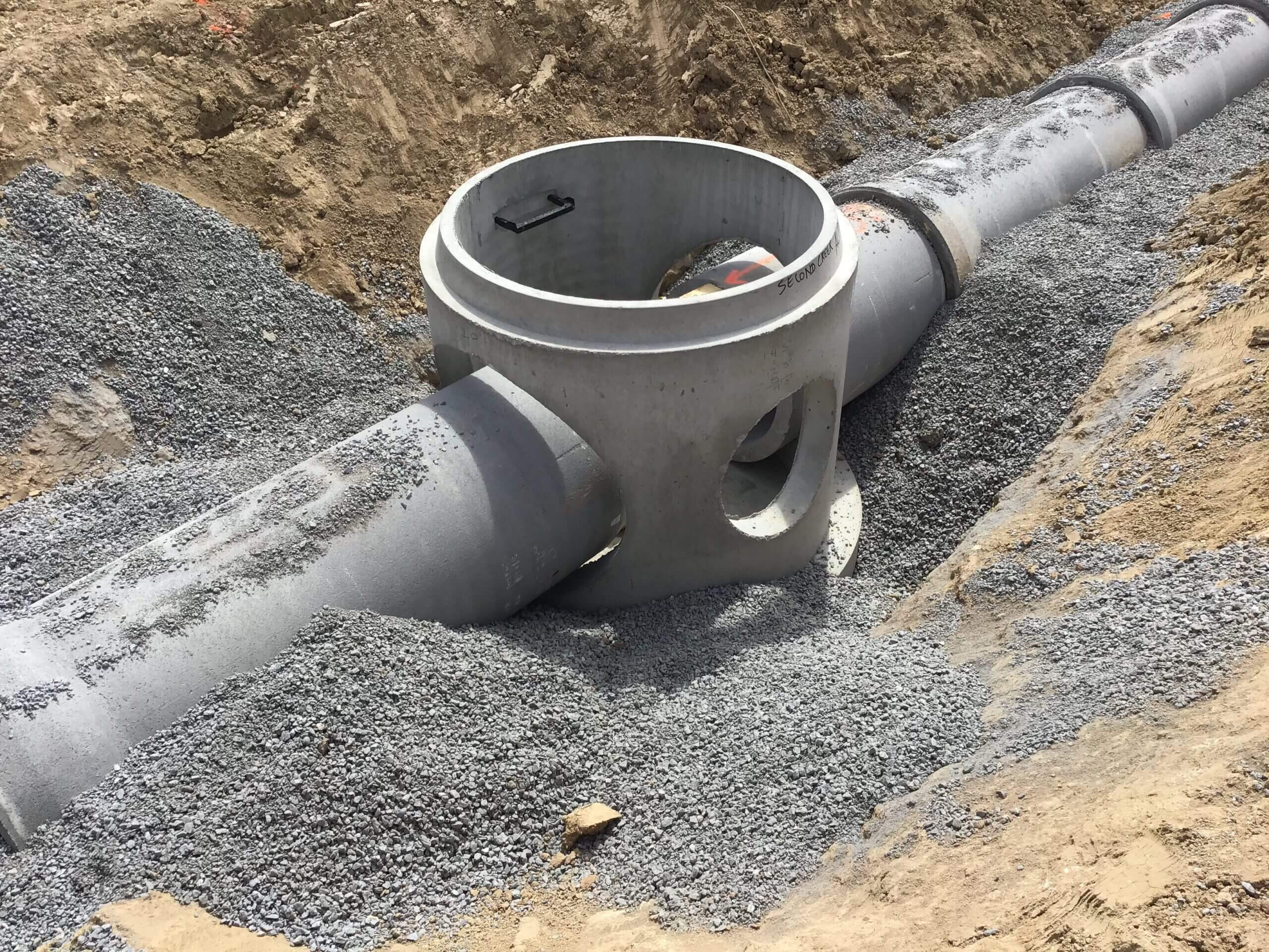 Precast concrete stormwater manhole installed between large drainage pipes in a gravel-lined trench at a construction site.