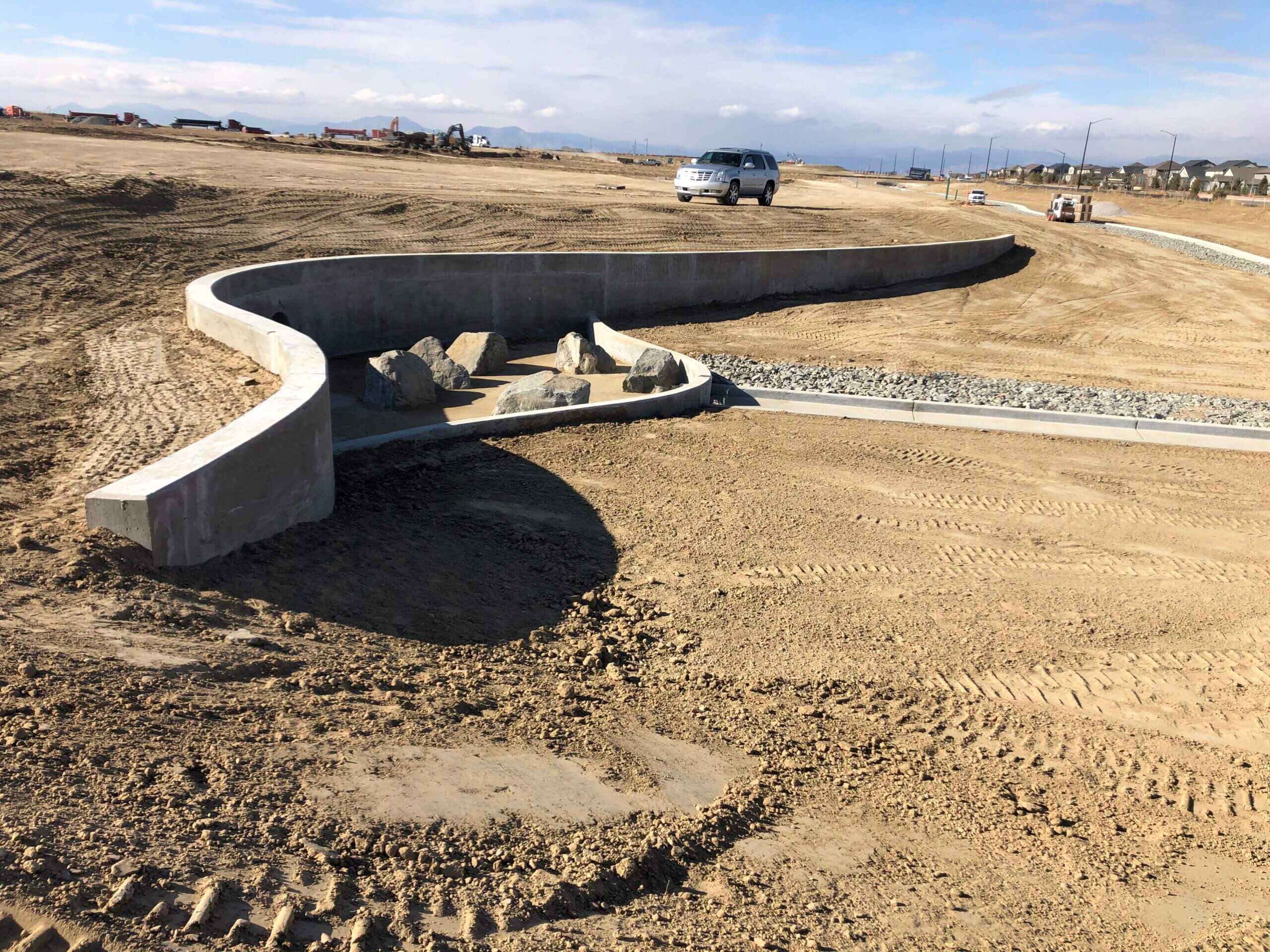 Curved concrete retaining wall and rock-lined drainage feature under construction in a large graded development site with mountains in the distance.