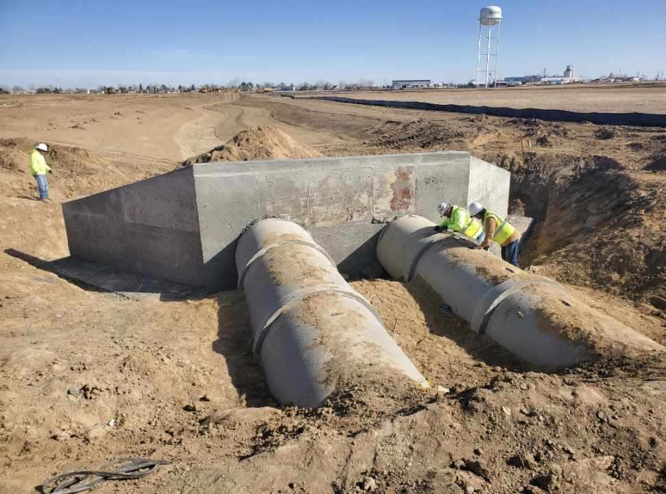 Workers install large concrete drainage pipes connected to a concrete culvert structure at an earthwork construction site with a water tower visible in the distance. Workers install large concrete drainage pipes connected to a concrete culvert structure at an earthwork construction site with a water tower visible in the distance.