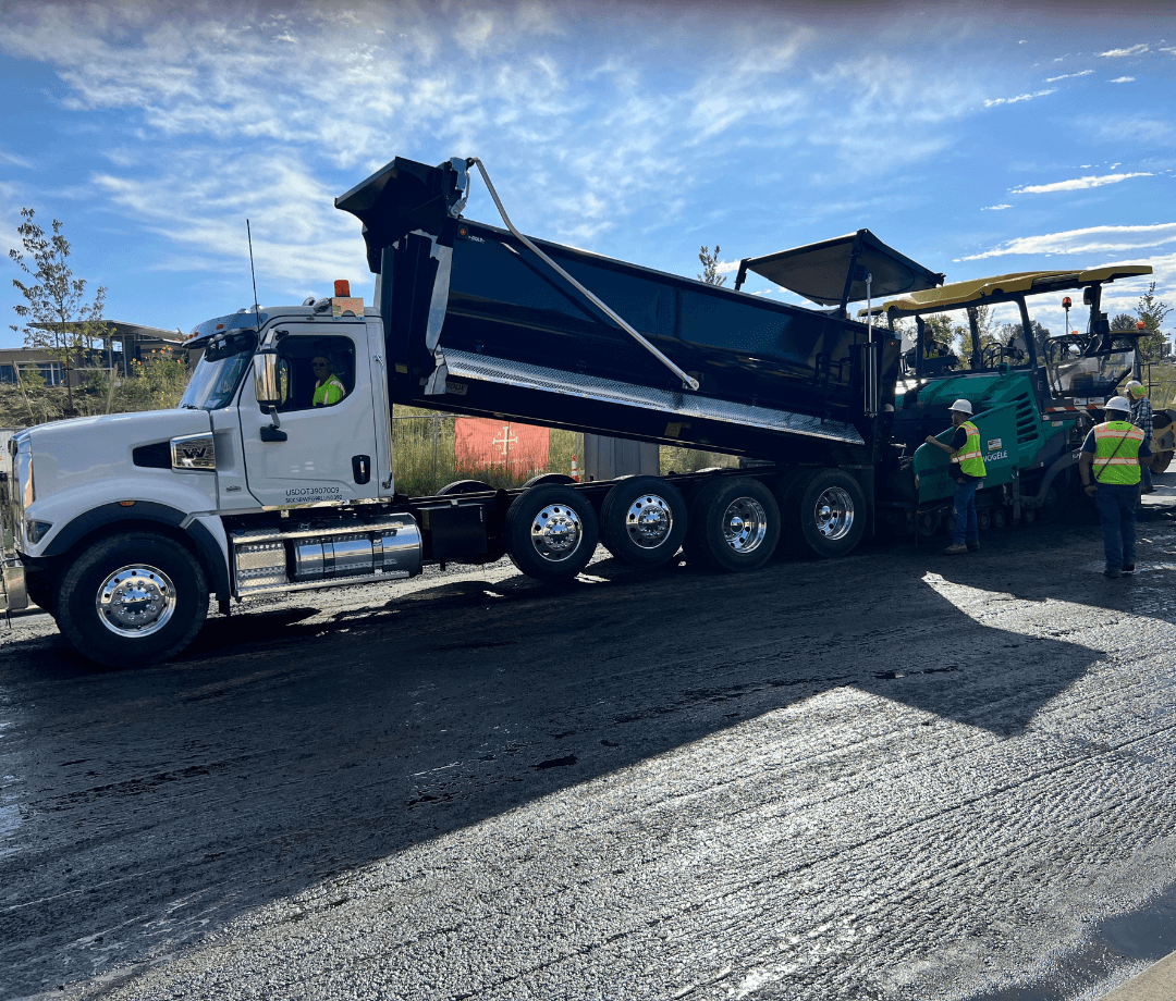 Dump truck unloading hot asphalt into a paving machine while construction workers monitor the paving operation on a roadway project. Dump truck unloading hot asphalt into a paving machine while construction workers monitor the paving operation on a roadway project.