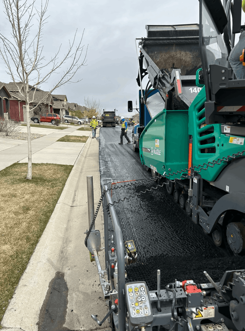 Asphalt paving machine laying fresh asphalt along a residential street while construction workers guide the paving process. Asphalt paving machine laying fresh asphalt along a residential street while construction workers guide the paving process.