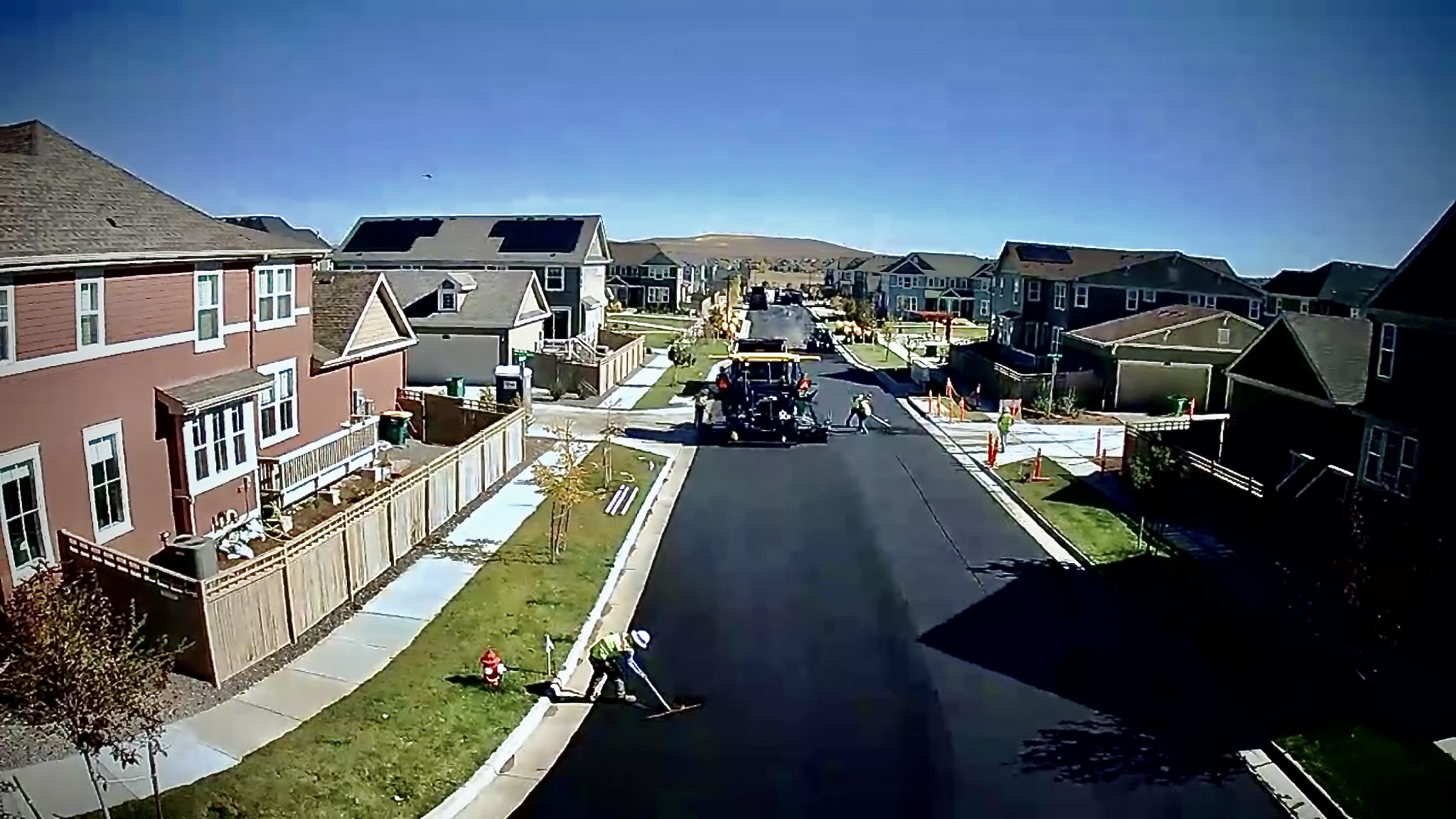 Road construction crew paving a freshly resurfaced residential street lined with homes and sidewalks.