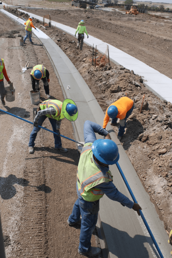 Construction workers wearing hard hats and safety vests install and finish a newly poured concrete curb and sidewalk along a roadside construction project. Construction workers wearing hard hats and safety vests install and finish a newly poured concrete curb and sidewalk along a roadside construction project.