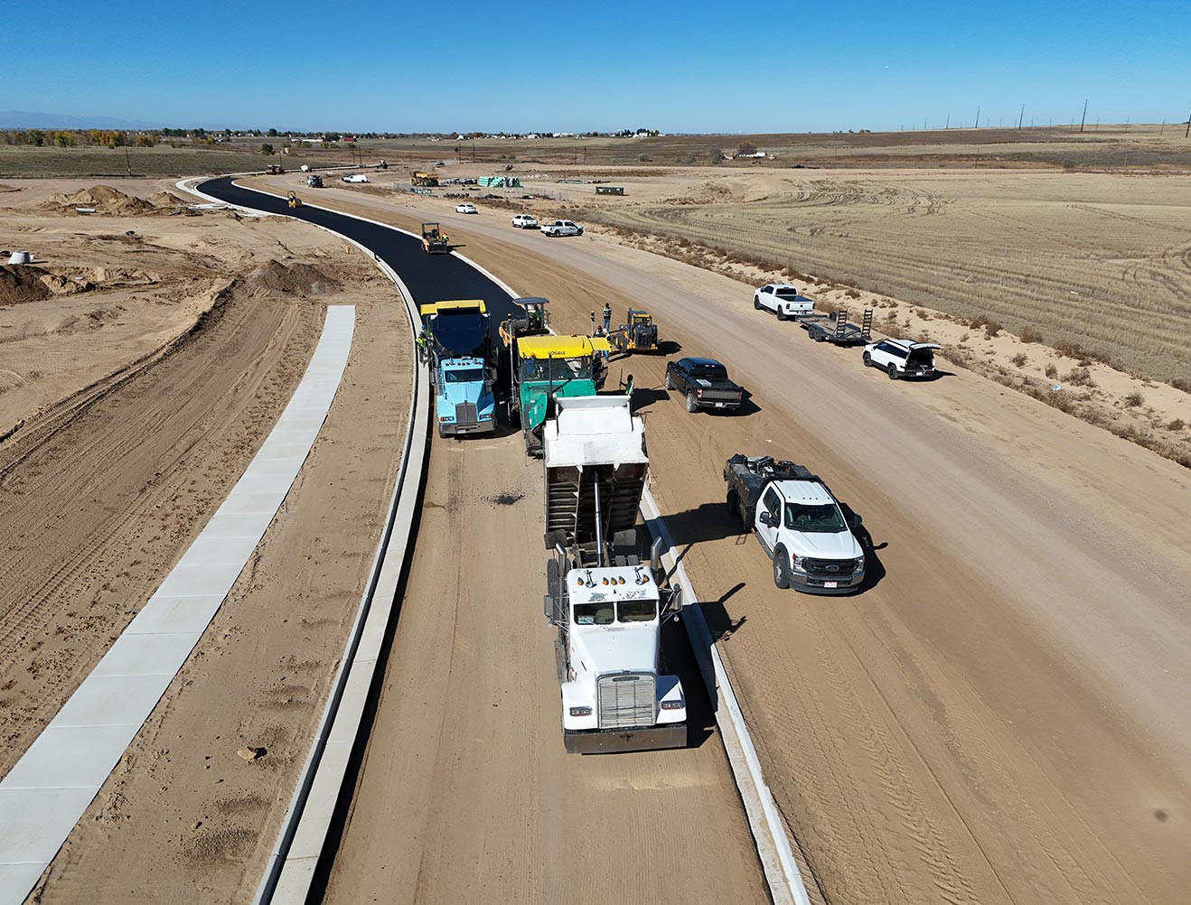 Aerial view of road construction crew paving fresh asphalt on a curved roadway with dump trucks and heavy equipment in an open rural area