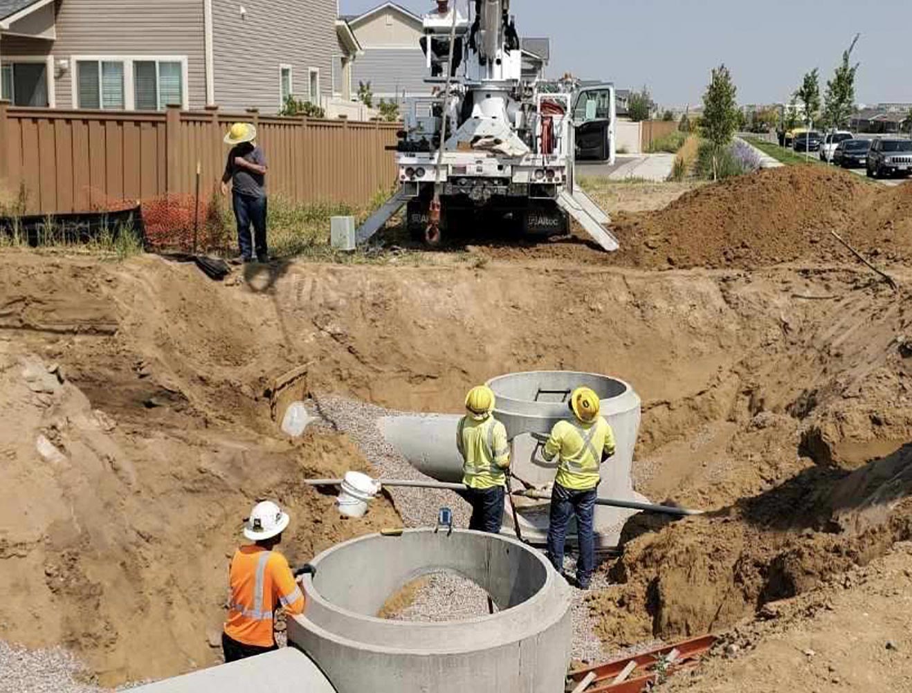 Construction workers in safety vests and hard hats installing precast concrete manhole sections in an excavated trench in a residential neighborhood